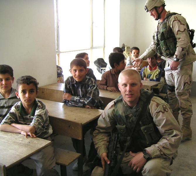 Photograph of Iraqi children in their class room smiling into the camera along with an American soldier who is visiting them. Photograph by an American soldier of C Co, 1/252 Army Reserve Battalion. 