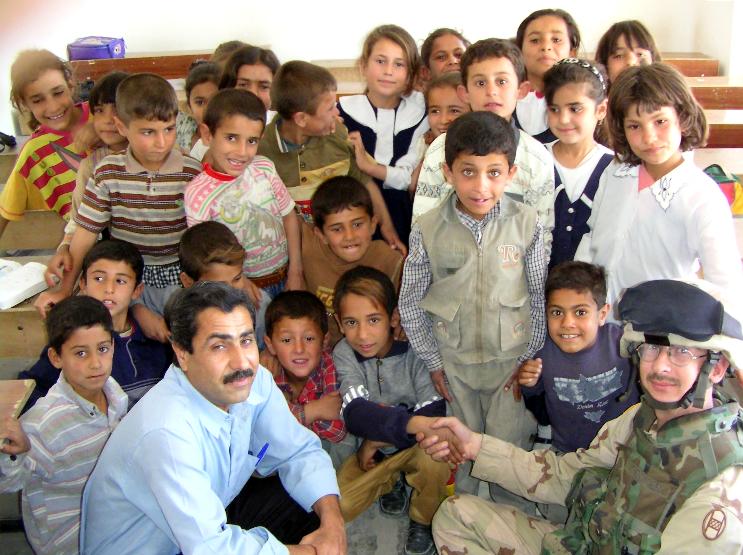 Photograph of Iraqi children in their class room with their teacher and an American soldier who is shaking the hand of one young boy. Photograph by an American soldier of C Co, 1/252 Army Reserve Battalion. 