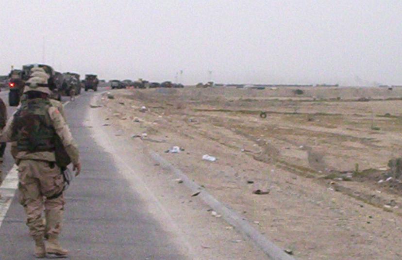 Photograph of a long line of military vehicles and men marching down a road in Iraq. Photograph by an American soldier of C Co, 1/252 Army Reserve Battalion. 